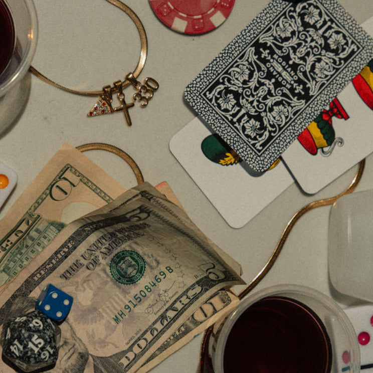 Tabletop game setup with cards, dice, money, and drinks on a surface. With the Youngstown Charm Necklace featuring charms. A pizza, a cross, and 330.